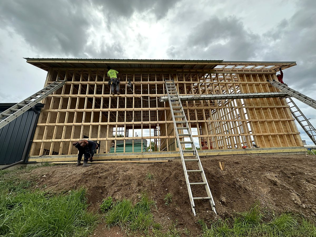 Large wood-framed building with roof on, crew members on ladders and scaffolding framing the wall studs, with black metal siding already on the neighboring wing