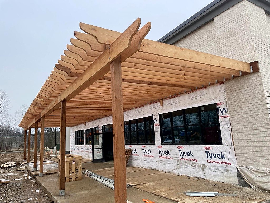 Timber pergola with scalloped rafters covering a walkway along a new commercial building wrapped in housewrap, with black storefront windows