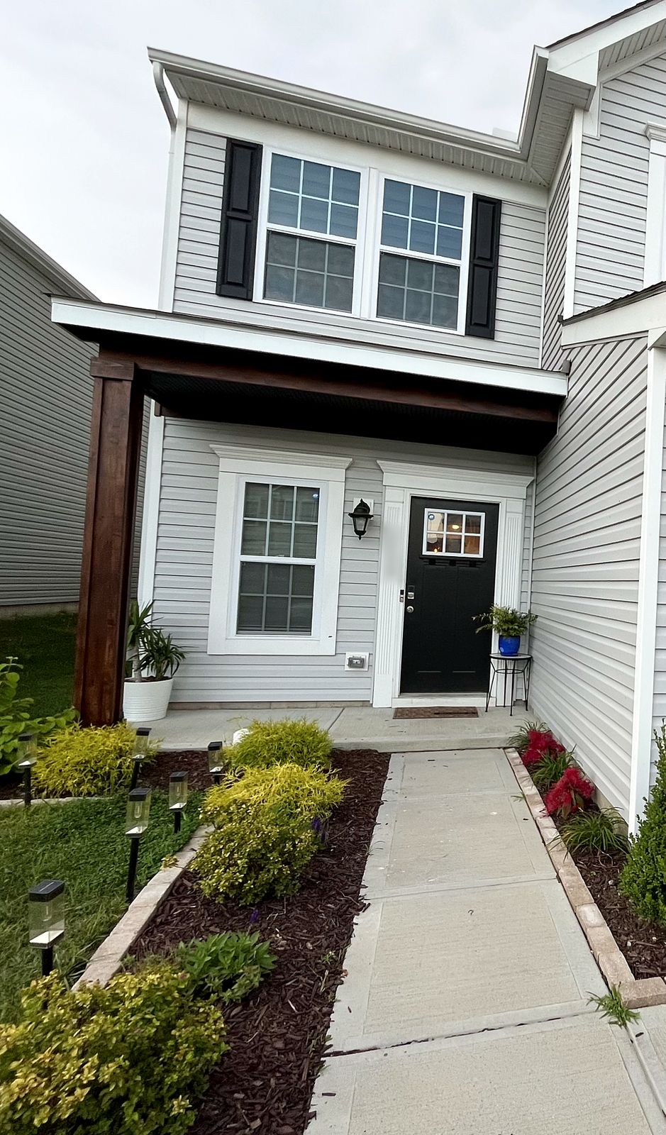 Finished front porch with a stained wood post and covered entry, a black front door and landscaped beds