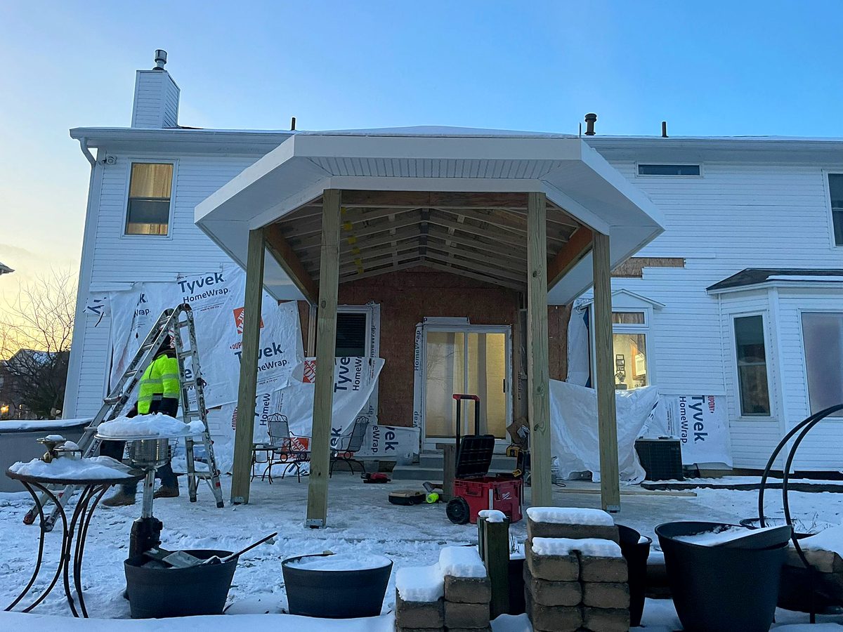 Covered patio addition under construction on a white two-story house, with pressure-treated posts, roof framing, white trim and housewrap, and a crew member beside a ladder