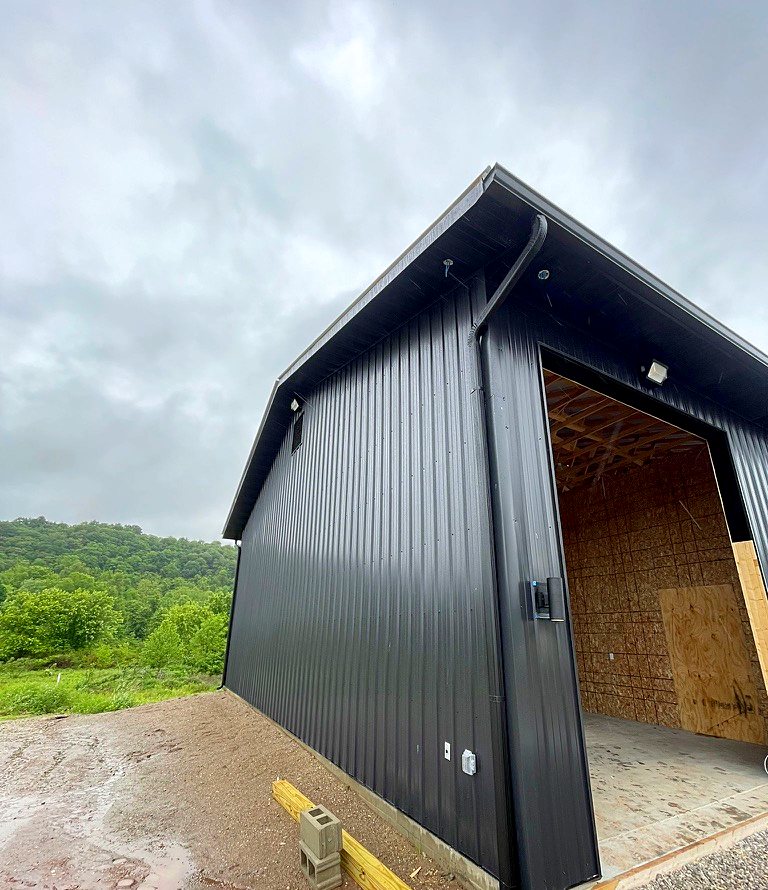Black metal building with a large open bay, roof edge and downspout, on a cloudy day with green hills behind
