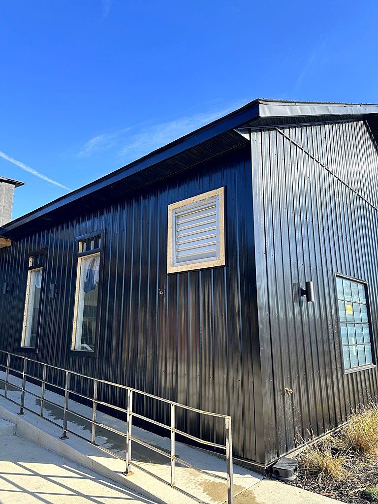 Black vertical metal siding on a commercial building under a clear blue sky, with windows, a louvered vent, wall lights, a metal roof edge and a handrail