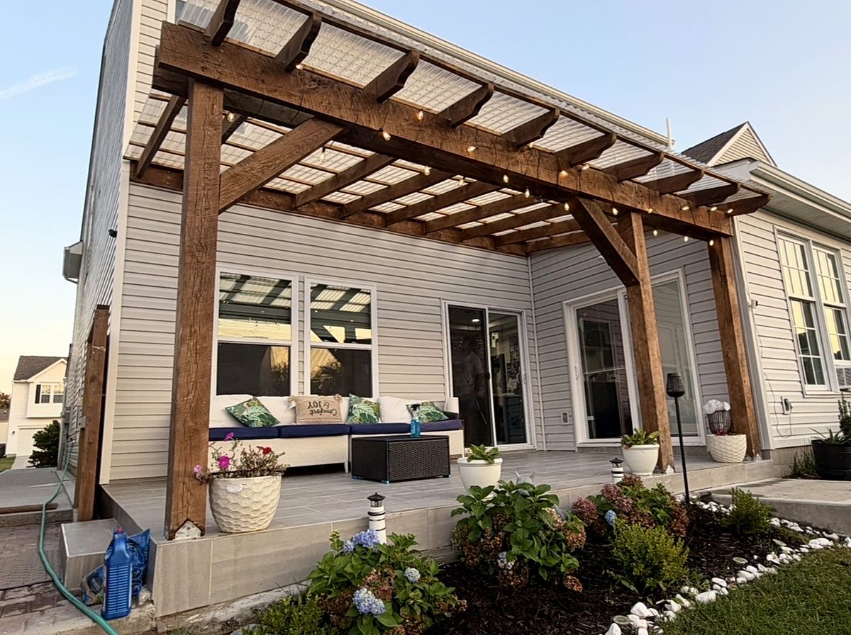 Stained timber pergola with clear roof panels over a tiled patio, with outdoor seating and flower beds