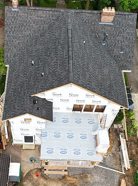 Aerial view of a house with a new dark architectural shingle roof, a rear addition wrapped in housewrap, and a dumpster in the driveway