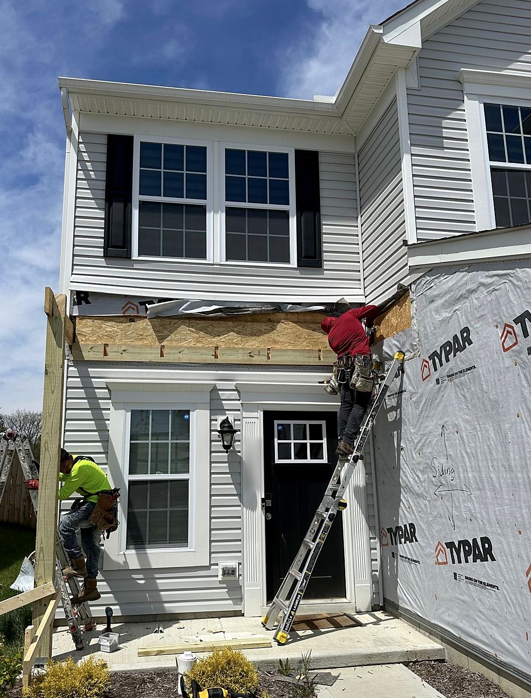 Two crew members on ladders framing a new porch header on a two-story home, with housewrap already on the side wall
